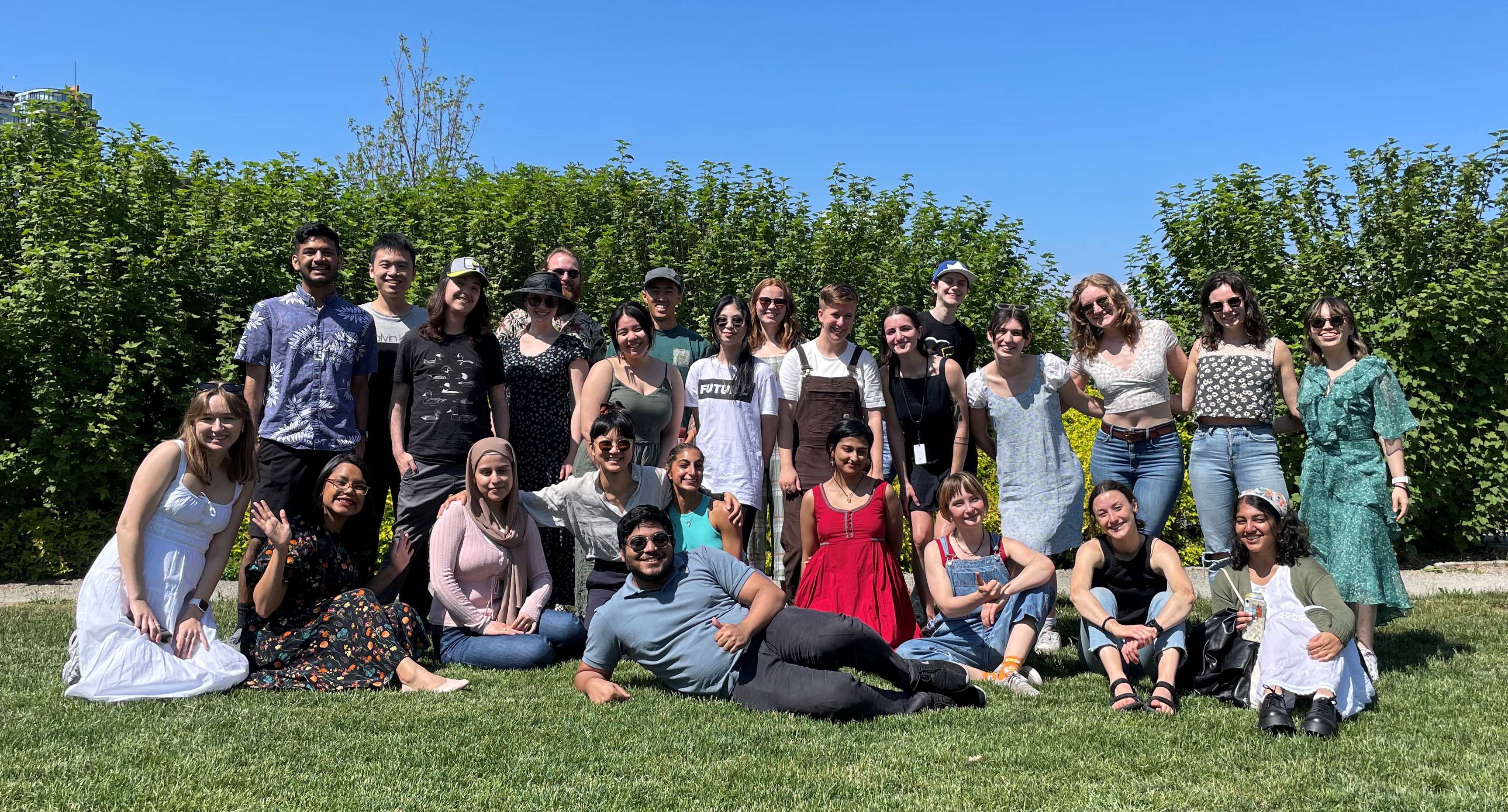 Large group of youth smiling, some sitting on the grass, some standing. In the background are green bushes against a bright blue sky