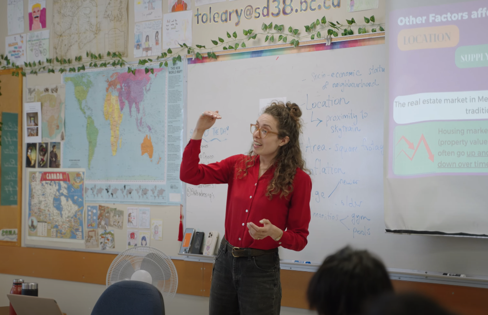 Photo of woman in red shirt speaking animatedly at the front of a classroom. 